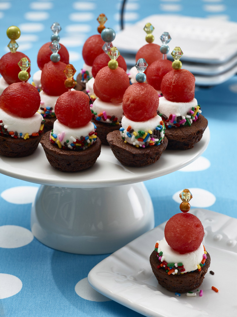 Dozen brownie bites set on white cake plate topped with decorative toothpicks. One brownie bite served on small dessert plate with more serving plates in background. Setting is aqua with white polka-dot tablecloth.