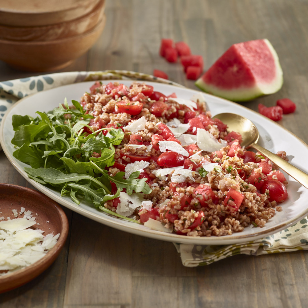 Plated salad on a white oval dish with arugula garnish. Pecorino Romana cheese on clay dish on side with wooden serving bowls and watermelon wedge and chunks in background. Setting is on a wooden table.