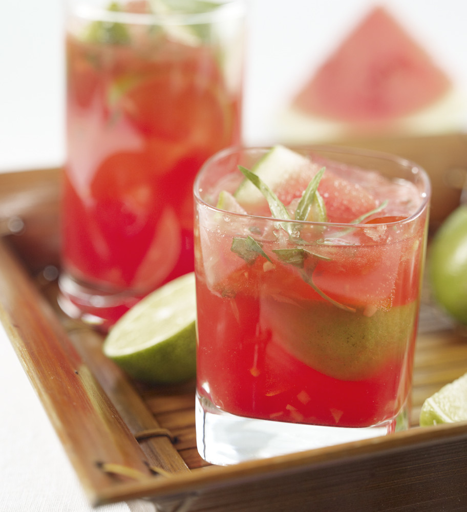 Two Watermelon Caipirinhas served in bamboo serving tray garnished with tarragon leaves and lime. Watermelon wedge in background.