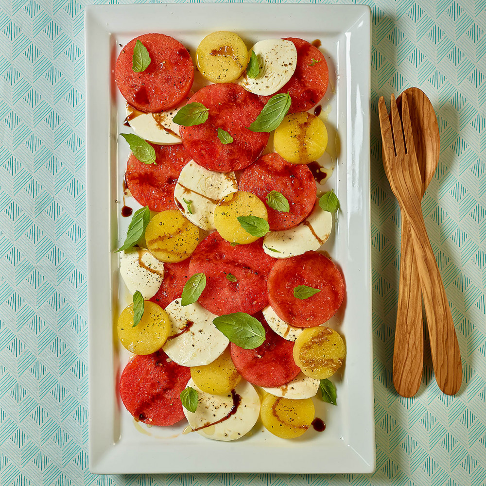Caprese salad served on a white rectangular plate with round slices of watermelon (red and yellow), mozzarella, garnished with basil and drizzled with balsamic vinegar. Plate is on an aqua patterned aqua and white tablecloth.