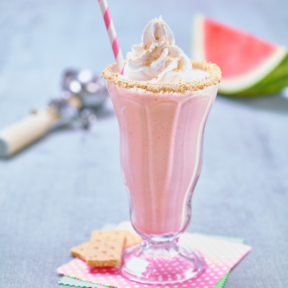 Watermelon Cheesecake Milkshake served in old-fashioned clear milkshake glass with pink and white straw. Garnished with graham cracker crumbs on rim, topped with whipped topping. Wedge of watermelon and ice cream scoop in background.