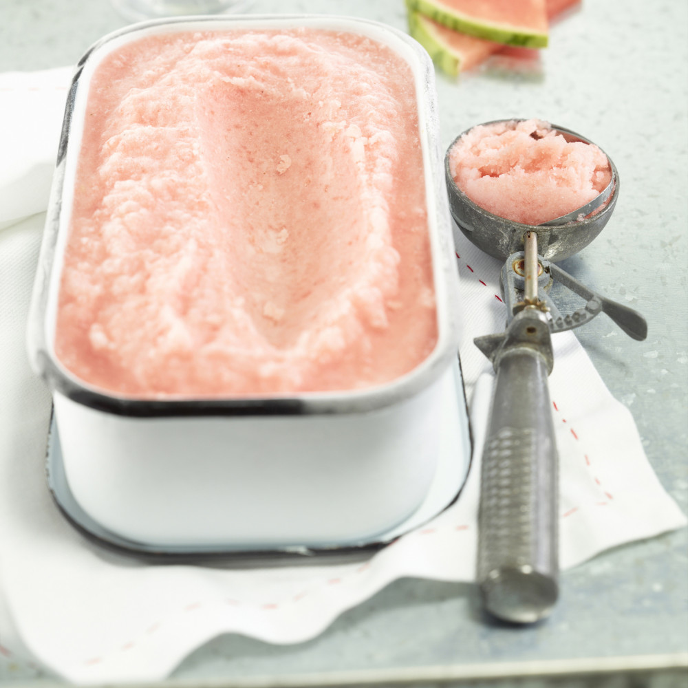 Creamy Watermelon Sherbet in freezer container with ice cream scoop on side with two dessert cups in background with two triangular pieces of watermelon.