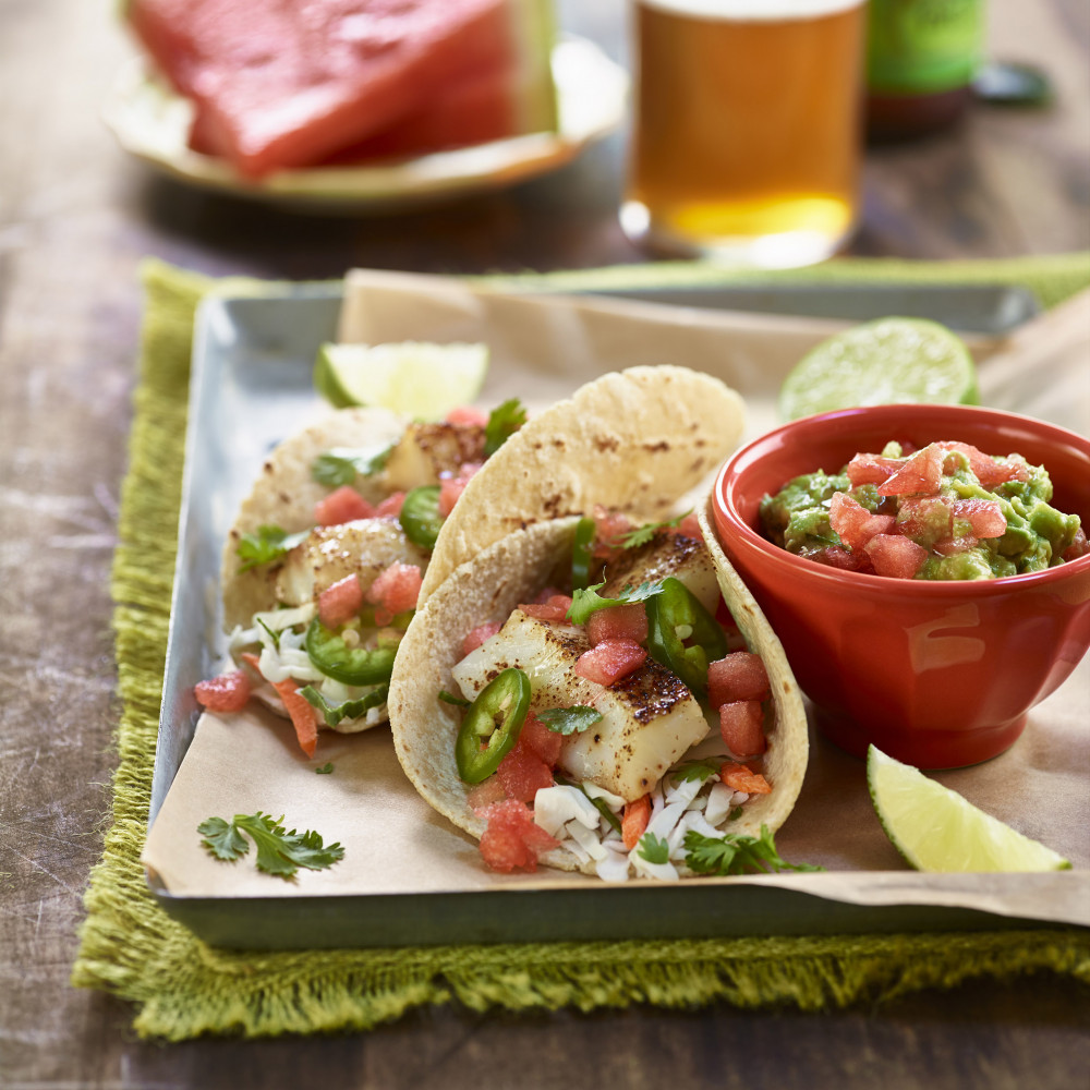 Watermelon fish tacos serving alongside bowl of watermelon guacamole. Watermelon wedges and clear glass of beer and beer bottle in background.