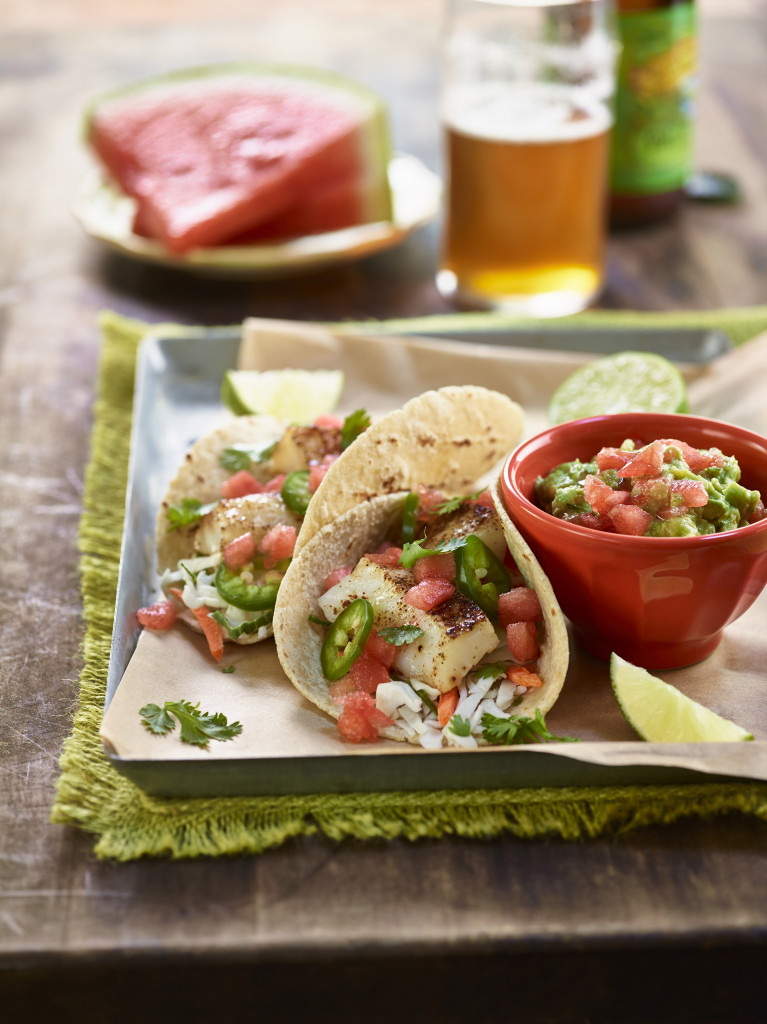Watermelon fish tacos serving alongside bowl of watermelon guacamole. Watermelon wedges and clear glass of beer and beer bottle in background.