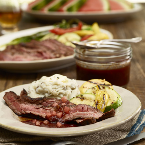 Flank-Steak with watermelon bourbon glaze plated on ivory stoneware with mashed potatoes and squash. Jar of bourbon glaze on side and additional plate and plate of watermelon wedges in background.