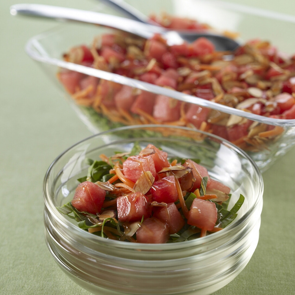 Singularly served salad on side of large serving bowl with serving spoons, clear bowls atop green tablecloth.