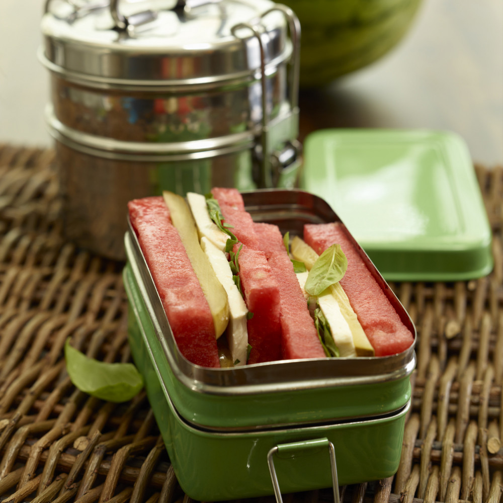 green tin lunchbox atop wicker mat with round-stacked lunchbox with recipe contents sliced and wedged in, basil garnish, whole watermelon in background