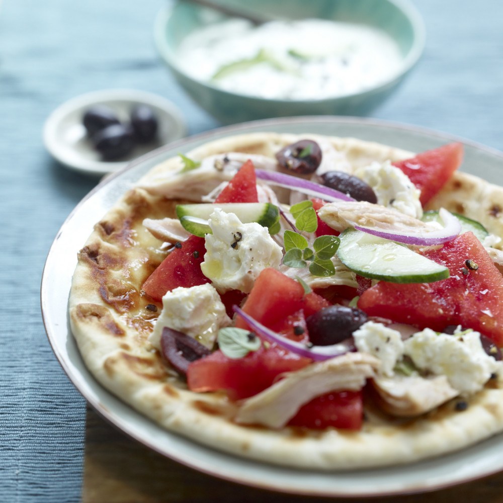 blue tablecloth, wooden cutting board with plated flatbread with tzatziki sauce and black olives in background