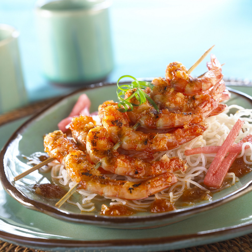 Grilled shrimp skewers served over noodles on stoneware. Garnished with watermelon stix.