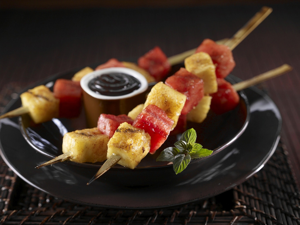 Three Grilled Watermelon Pineapple Skewers with ramekin of warm fudge sauce set on black plate. Sprig of mint leaves as garnish.