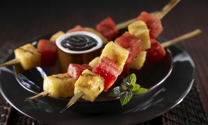 Three Grilled Watermelon Pineapple Skewers with ramekin of warm fudge sauce set on black plate. Sprig of mint leaves as garnish.