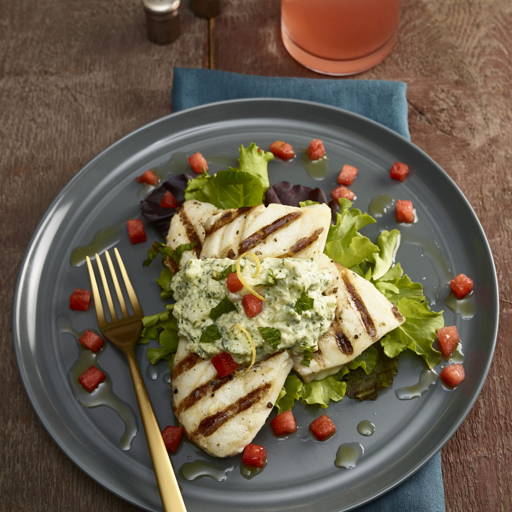 Halibut with Tzatziki sauce and diced watermelon plated on teal blue stoneware plate on teal blue cloth napkin with gold-toned fork. Small salt and pepper shakers on the side along with a clear glass of iced aqua-fresca. Background is wooden table.