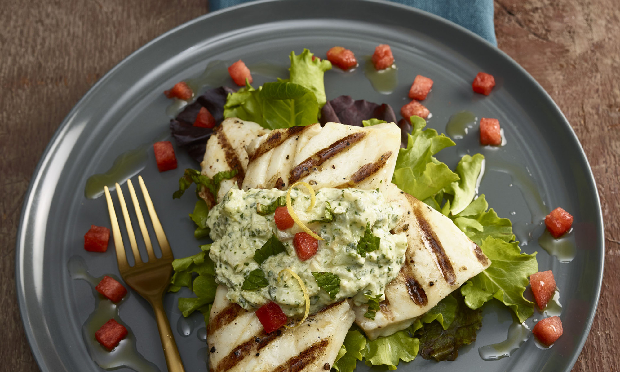 Halibut with Tzatziki sauce and diced watermelon plated on teal blue stoneware plate on teal blue cloth napkin with gold-toned fork. Small salt and pepper shakers on the side along with a clear glass of iced aqua-fresca. Background is wooden table.