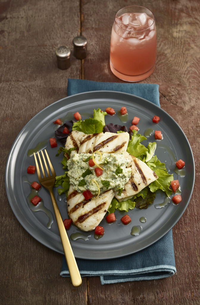 Halibut with Tzatziki sauce and diced watermelon plated on teal blue stoneware plate on teal blue cloth napkin with gold-toned fork. Small salt and pepper shakers on the side along with a clear glass of iced aqua-fresca. Background is wooden table.