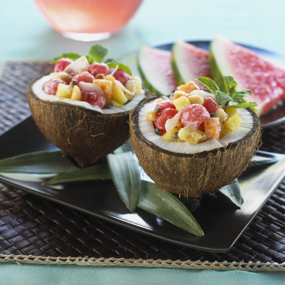 Watermelon Hawaiian Salad served in coconut halves on a black square plate on black woven placemat. Watermelon triangular slices on side and clear pitcher of aqua fresca in background.