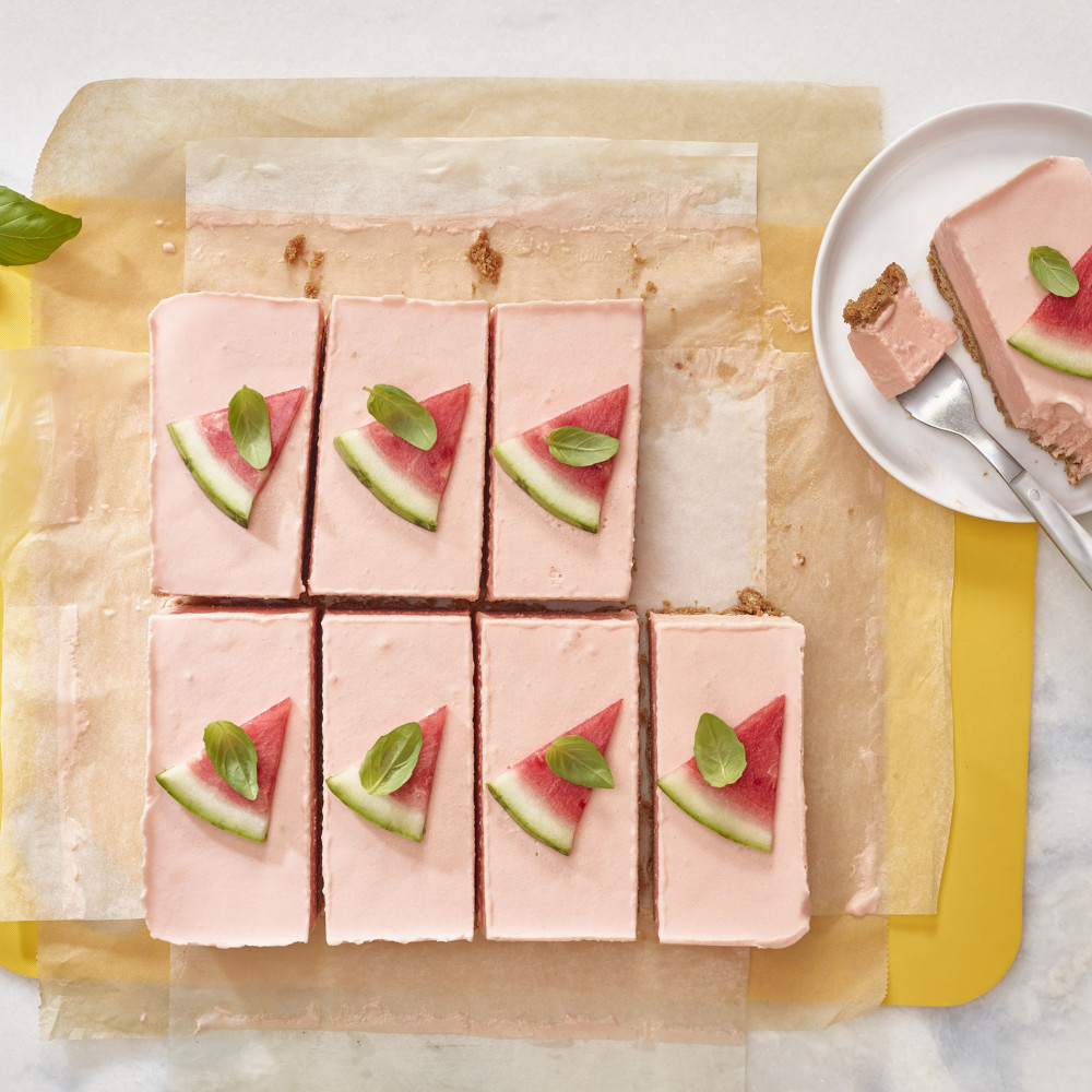 Watermelon Ice Cream Bars set on a yellow cutting board lined with parchment paper. One bar on plate with fork, seven pieces cut, garnished with triangular cut watermelon and small leaf of basil.