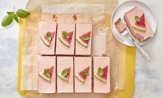 Watermelon Ice Cream Bars set on a yellow cutting board lined with parchment paper. One bar on plate with fork, seven pieces cut, garnished with triangular cut watermelon and small leaf of basil.