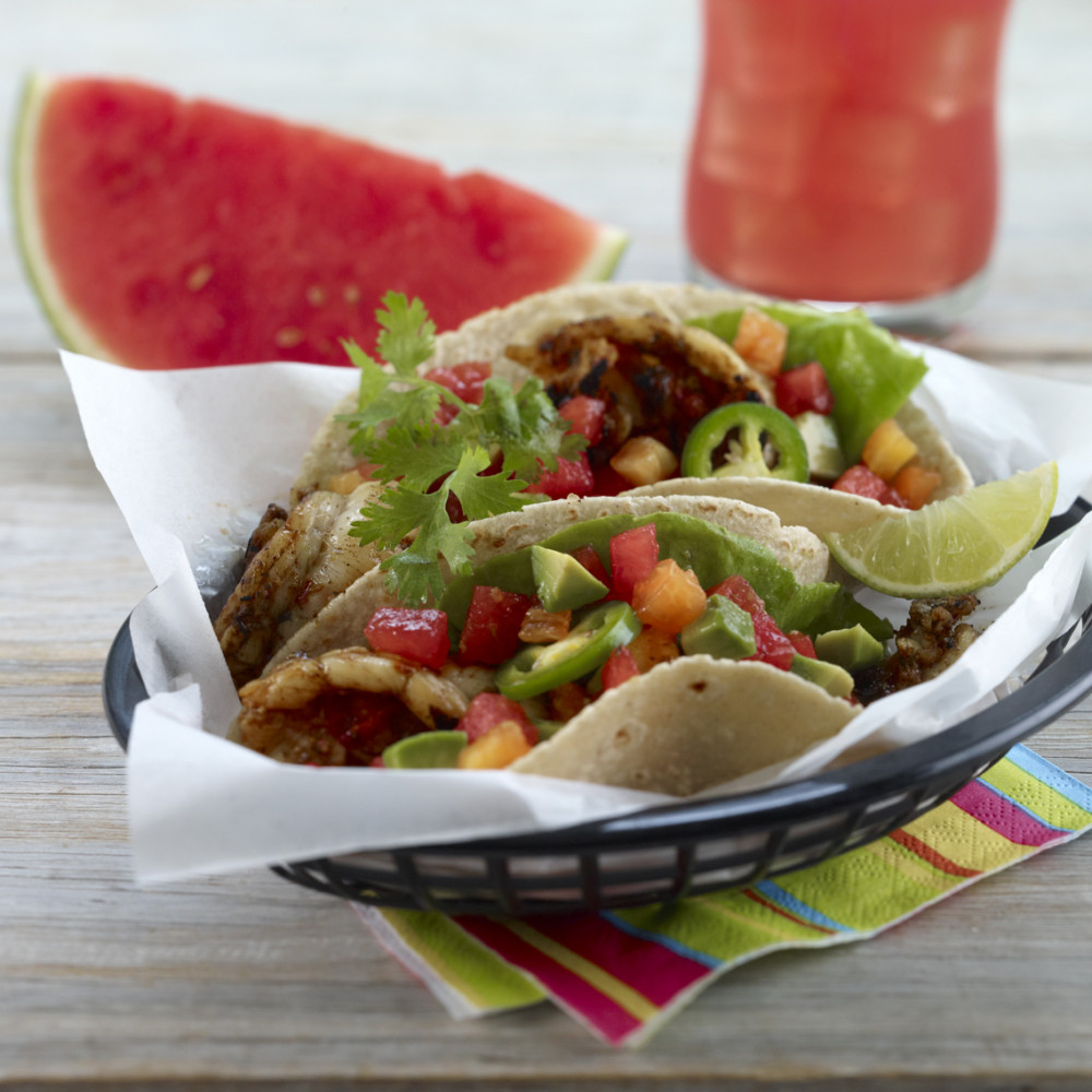 Tacos in a black serving basket with food-grade tissue on a colorful striped paper napkin on wooden table. Watermelon wedge and watermelon agua-fresca in background.
