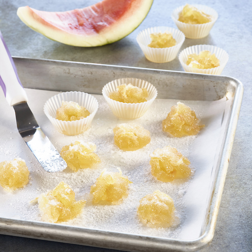 Lemon-Laced Rind Candy on baking sheet, some in mini paper cups. Watermelon rind wedge in background, without flesh.