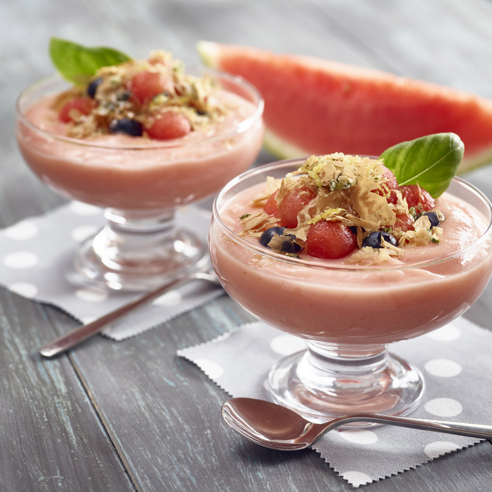 Two clear dessert cups with servings of Lemon Basil Crunch Watermelon Custard garnished with basil leaves. Watermelon wedge in background.