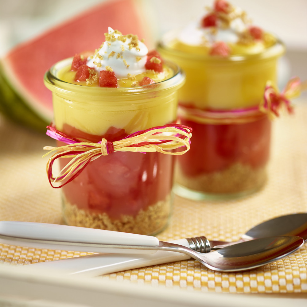 Watermelon Lemonade Cream Pie Shooters in two small clear jars with layered ingredients. Jars wrapped with raffia/bow. Topped with diced watermelon and graham cracker crumbs. Watermelon wedge in background.
