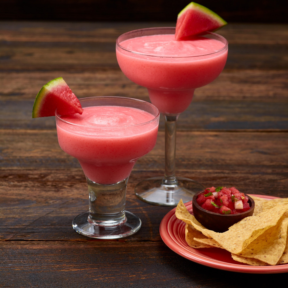 Two margaritas garnished with watermelon triangular cuts. Small plate of chips and Fire & Ice Salsa shown in foreground.