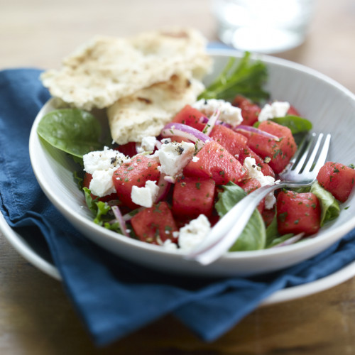Watermelon Mediterranean Salad in a white bowl atop a white plate with blue napkin. Pita bread on side of salad.