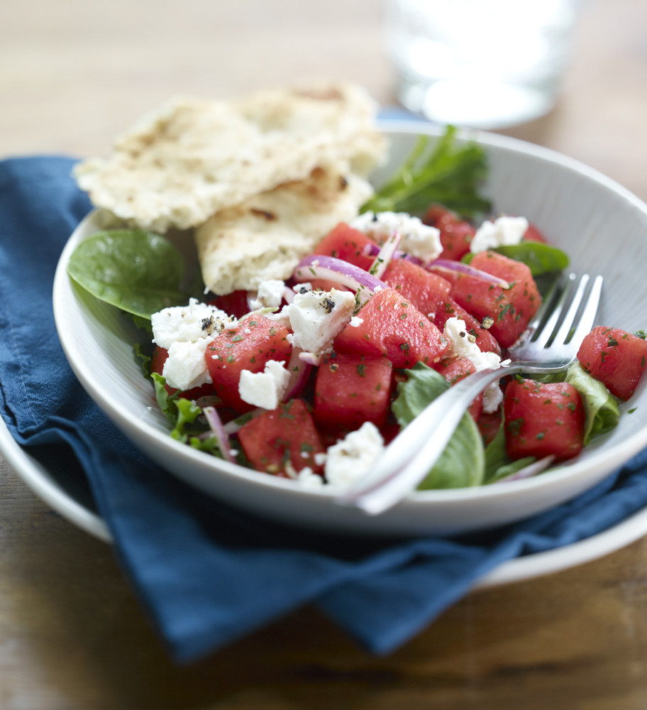 Watermelon Mediterranean Salad in a white bowl atop a white plate with blue napkin. Pita bread on side of salad.