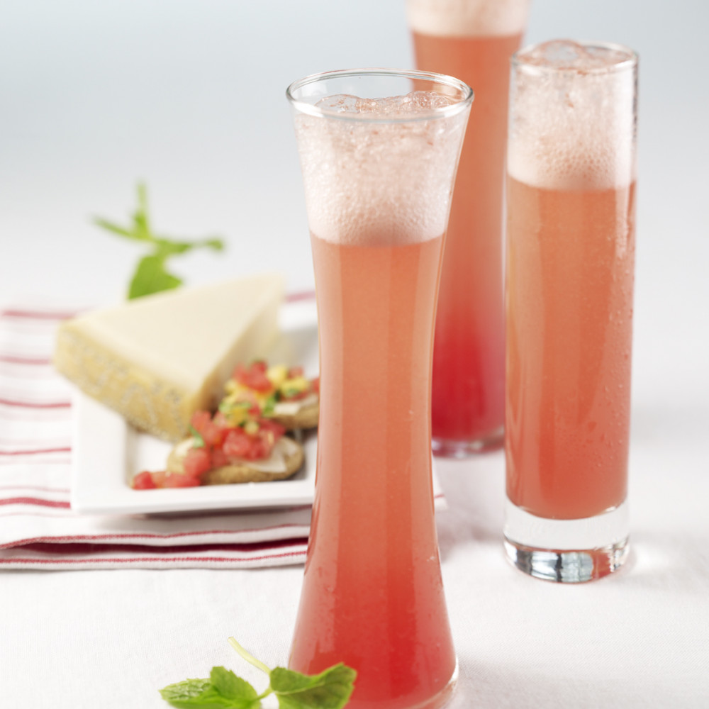 Three Watermelon Mimosas served in tall fluted clear glasses. Sprig of mint leaves on side. Plate of appetizer in background.