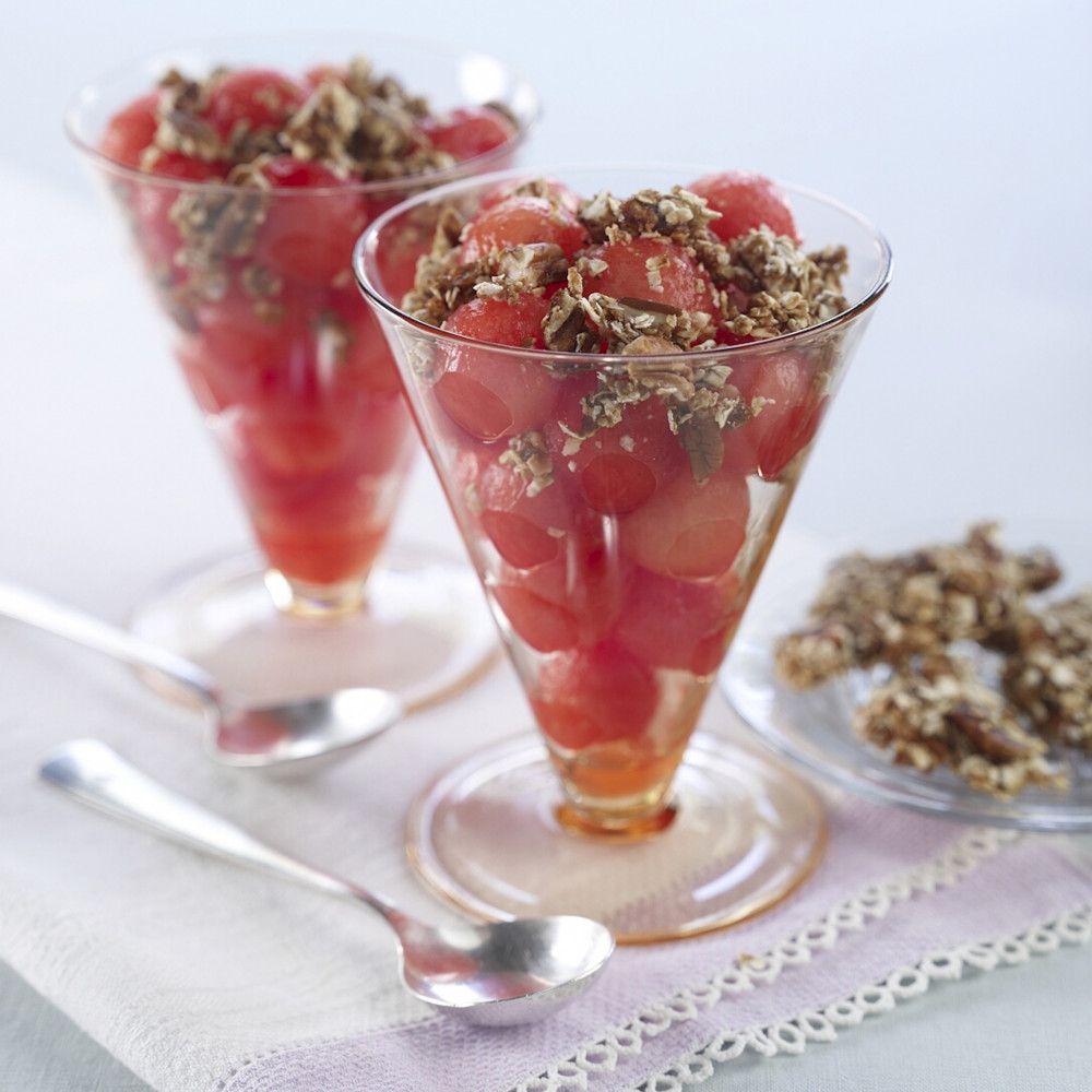 Two parfait glasses filled with watermelon balls and topped with oat crumble. Oat crumble in background.