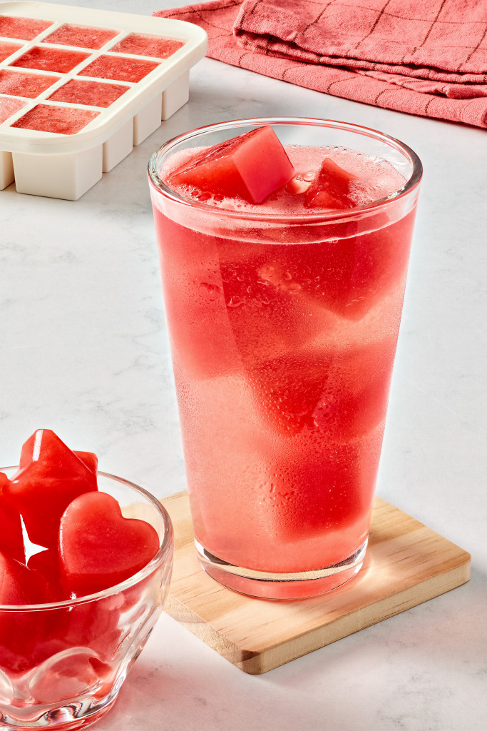watermelon ice cubes floating in a clear glass. A filled ice cube tray is in the background.