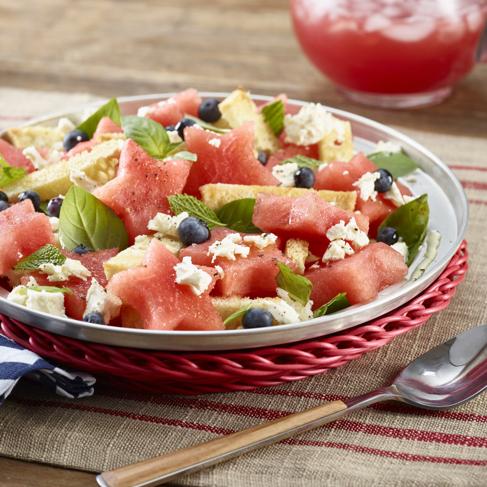 Panzanella Watermelon Salad plated on a red wicker charger. Pitcher of iced watermelon aqua fresca in background.