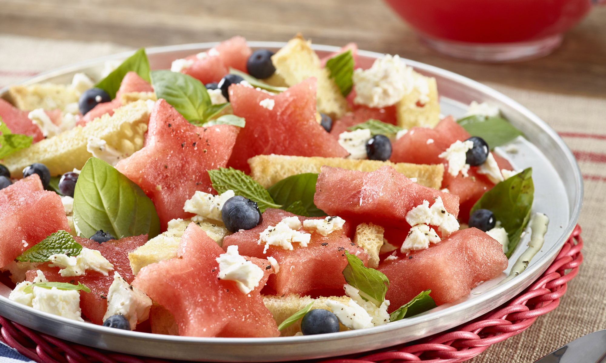 Panzanella Watermelon Salad plated on a red wicker charger. Pitcher of iced watermelon aqua fresca in background.