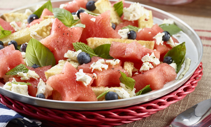 Panzanella Watermelon Salad plated on a red wicker charger. Pitcher of iced watermelon aqua fresca in background.