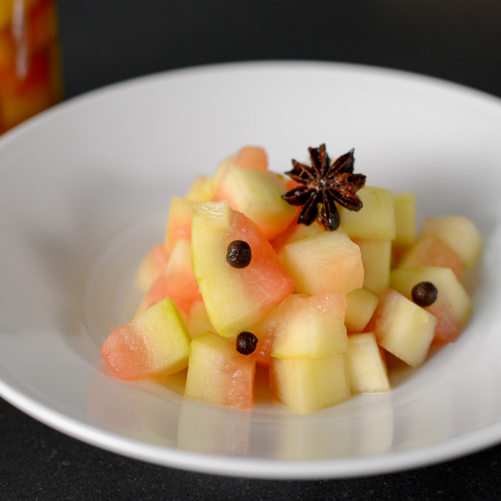 Pickled Watermelon Rind in white wide-rimmed bowl on black countertop. Garnished with allspice berries and star anise pod. Clear jar of Rind in background.