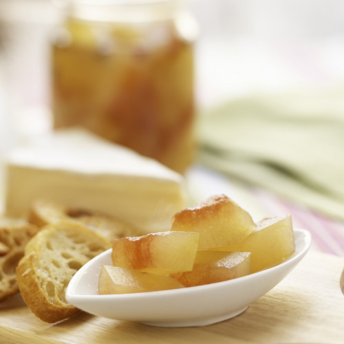 Pickled watermelon rind served on small condiment dish. Bread slices, cheese and jar of pickled rind in background, on wooden cutting board