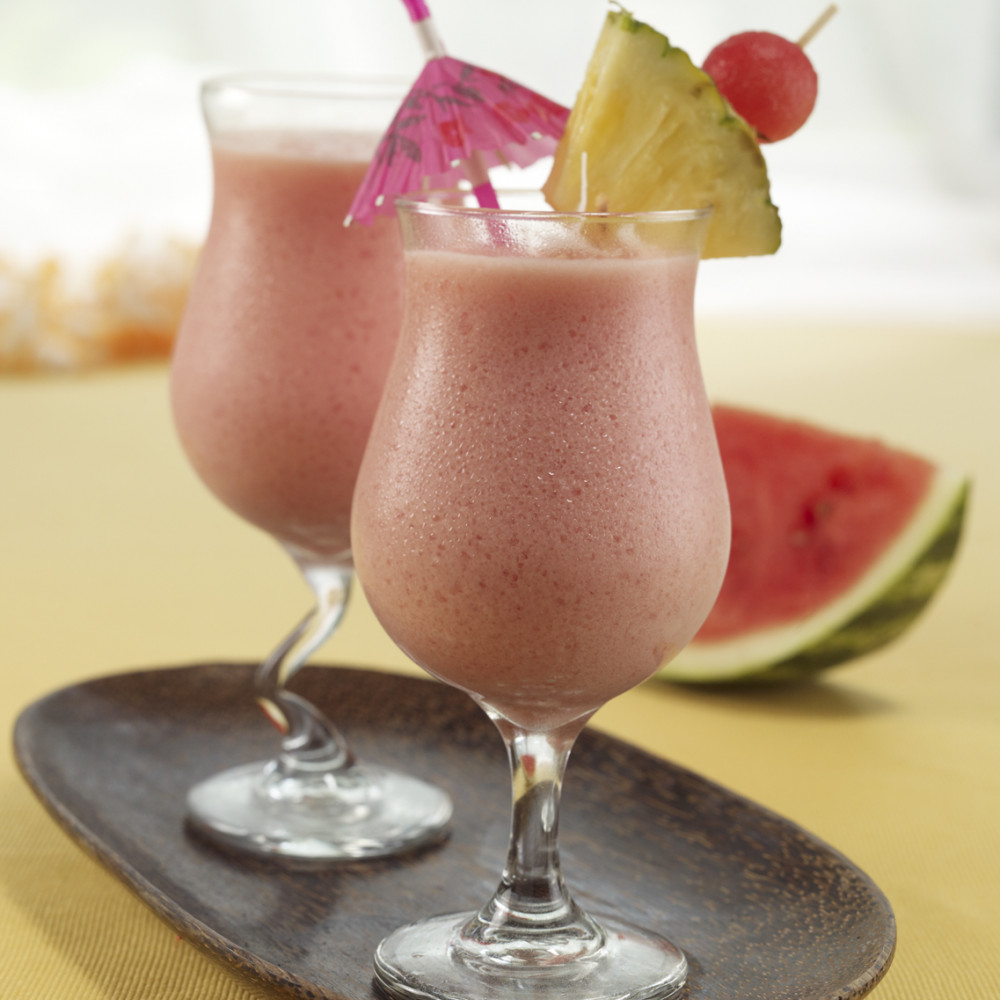 Two watermelon piña coladas served in clear cocktail glasses garnished with skewered pineapple and watermelon ball. Decorative straw. Watermelon wedge in background.