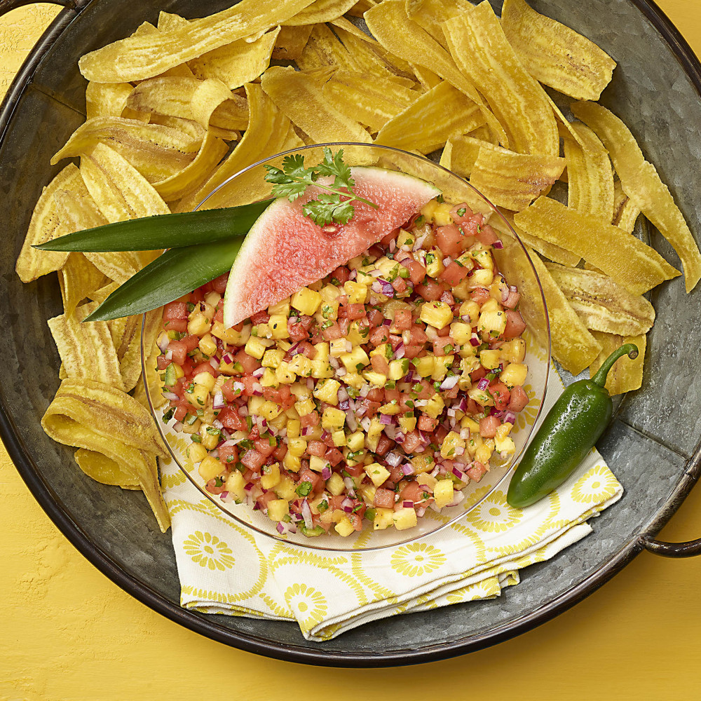 tray set with clear bowl of watermelon pineapple salsa with watermelon, jalapeño, cilantro and plantain green leaves as garnish with plantain chips alongside