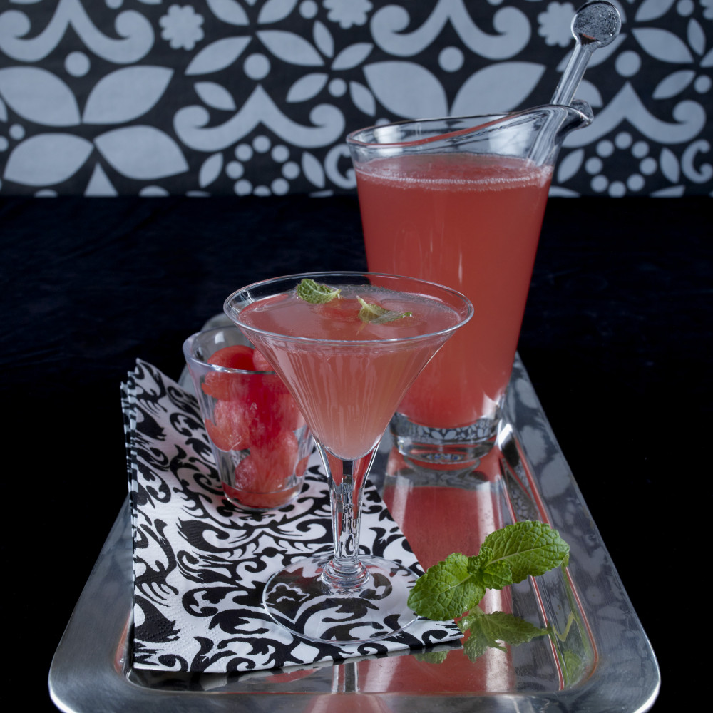 Pink Watermelon Chardonnay Sparkler served in martini glass with serving pitcher and small glass of watermelon balls. Garnished with mint leaves. Served on rectangular metal tray with black and white napkin and background.