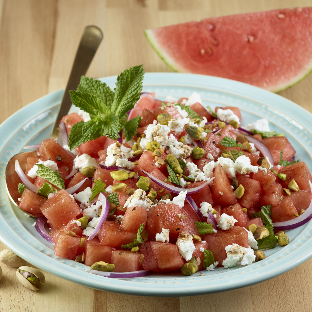 Watermelon & Pistachio Salad on a blue plate atop wooden cutting board or table with onion and mint as garnishes on top and pistachio (in shells) on wood, also watermelon wedge in background. Serving spoon on plate.
