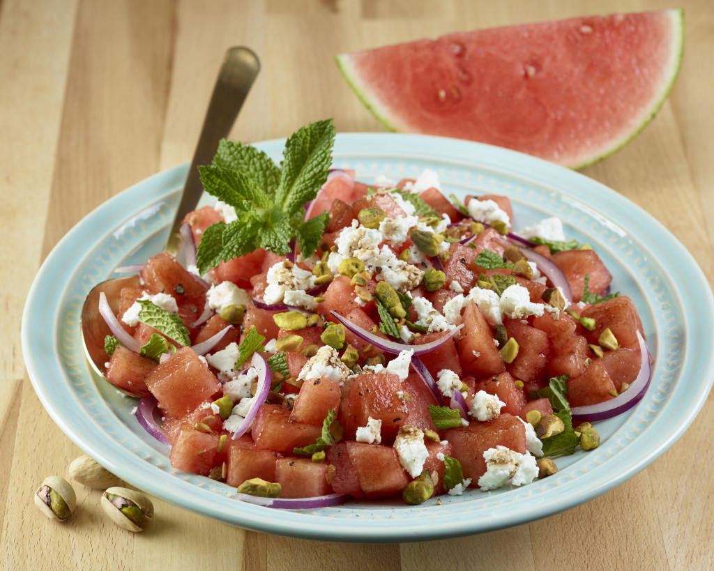 Watermelon & Pistachio Salad on a blue plate atop wooden cutting board or table with onion and mint as garnishes on top and pistachio (in shells) on wood, also watermelon wedge in background. Serving spoon on plate.
