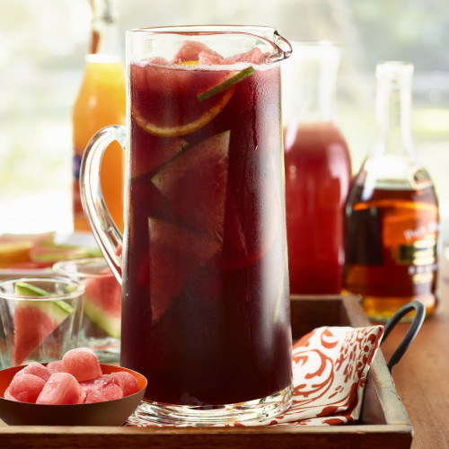 Tall, clear glass pitcher of Watermelon Pomegranate Sangria with cuts of watermelon & citrus in pitcher set in wooden tray. Partial bottles of some ingredients seen in background.