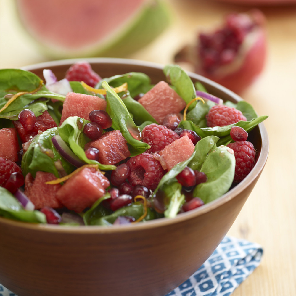 Watermelon Pomegranate Toss Salad in a wooden bowl atop a blue and white napkin with watermelon wedge and pomegranate halve in background.