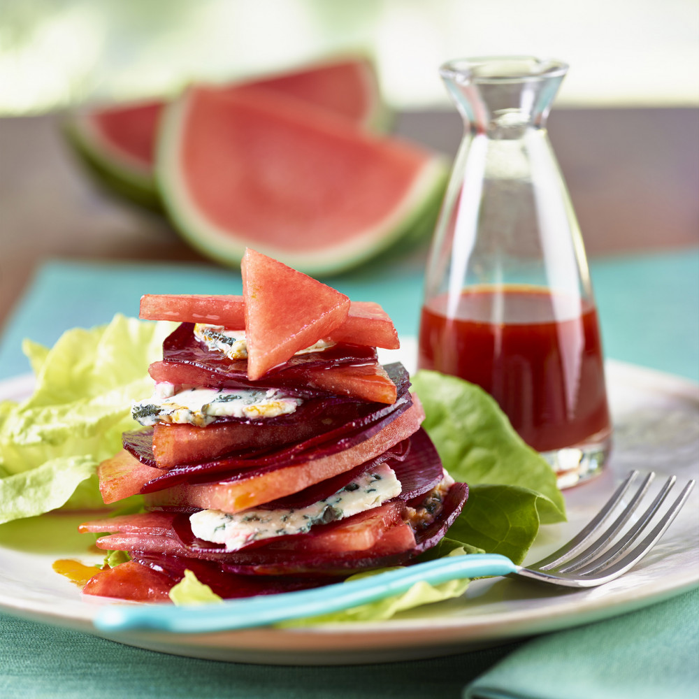 plated stacked watermelon red hot beet salad on turquoise napkin with fork and lettuce garnish with watermelon wedges in background. Carafe of dressing on side.