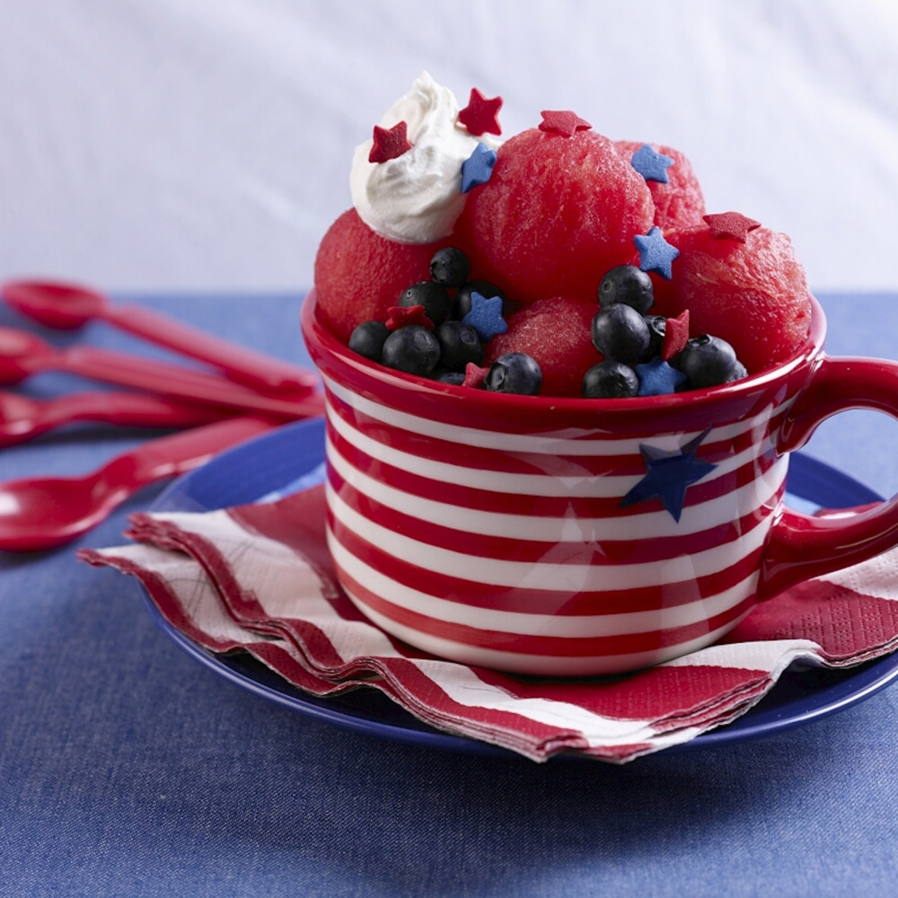 Sundae arranged in red white cup with blue star on a saucer. Red spoons in background.