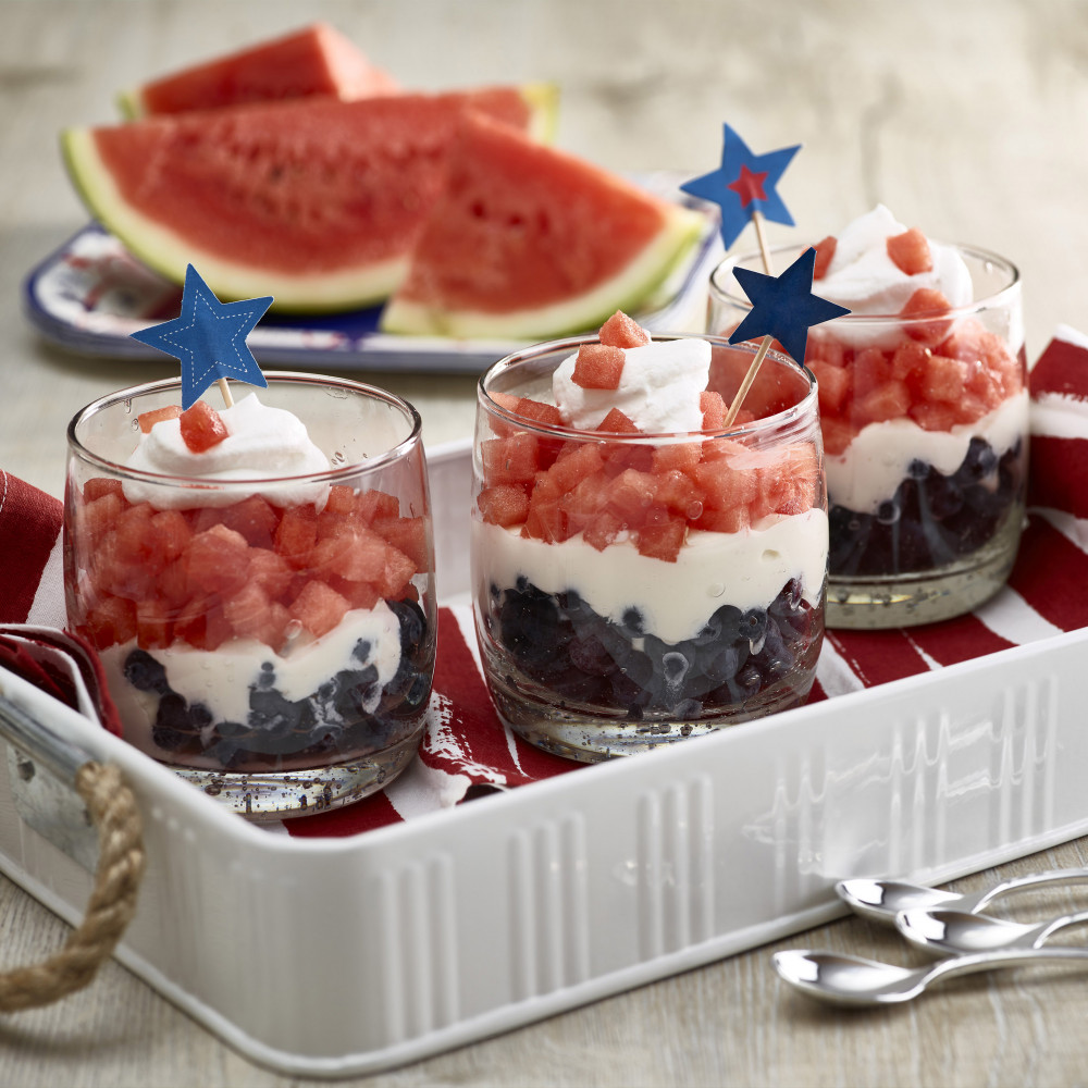 Three clear short glasses set in white serving tray. Glasses filled with layered blueberries, yogurt and diced watermelon topped with whipped cream and decorative toothpicks. Watermelon halves and wedges in background.