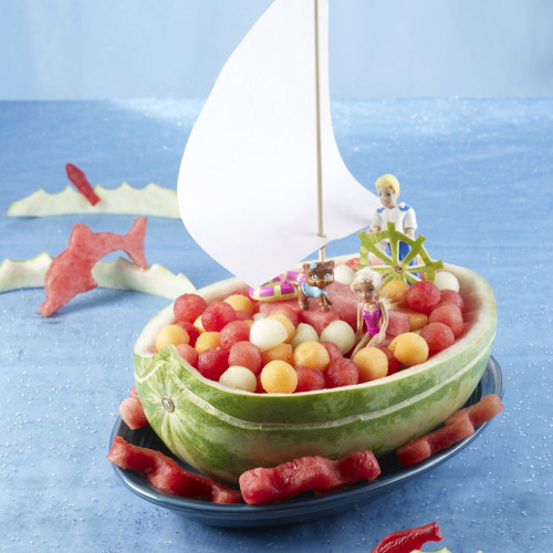 Sailboat set on large oval platter with watermelon balls on sides and inside. Setting on blue tablecloth. Fish-shaped cutouts on sides, sail in boat and captain at helm.