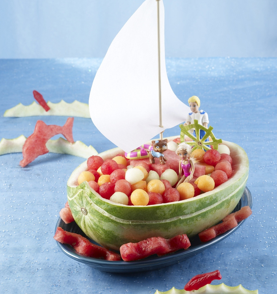 Sailboat set on large oval platter with watermelon balls on sides and inside. Setting on blue tablecloth. Fish-shaped cutouts on sides, sail in boat and captain at helm.