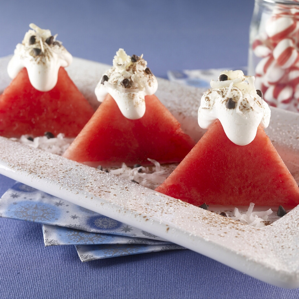 Three pyramid shaped watermelon cuts served on rectangular plate topped with whipped cream, shredded coconut, white chocolate shavings and mini chocolate chips. Jar of peppermint candies in background.