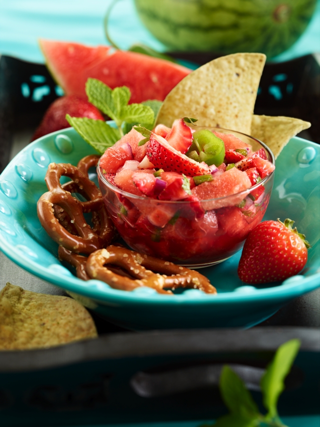 clear bowl with strawberry mint salsa set in turquoise bowl alongside pretzels, chips, whole strawberry as garnish and watermelon in background